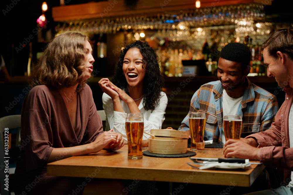 Happy young multiracial group of friends in casual clothing talking and laughing over dinner at bar
