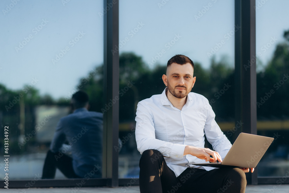 Young businessman working online with a laptop while sitting on a bench outside of his office building Young businessman working online with a laptop while sitting on a bench outside of his office building
