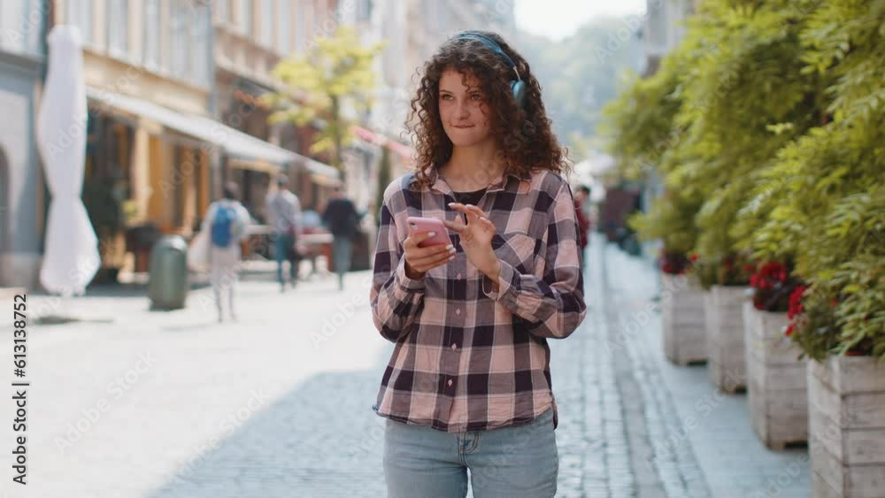Happy relaxed overjoyed young woman in wireless headphones choosing, listening favorite energetic disco rock n roll music in smartphone dancing outdoors. Girl walking in urban sunshine city street Happy relaxed overjoyed young woman in wireless headphones choosing, listening favorite energetic disco rock n roll music in smartphone dancing outdoors. Girl walking in urban sunshine city street