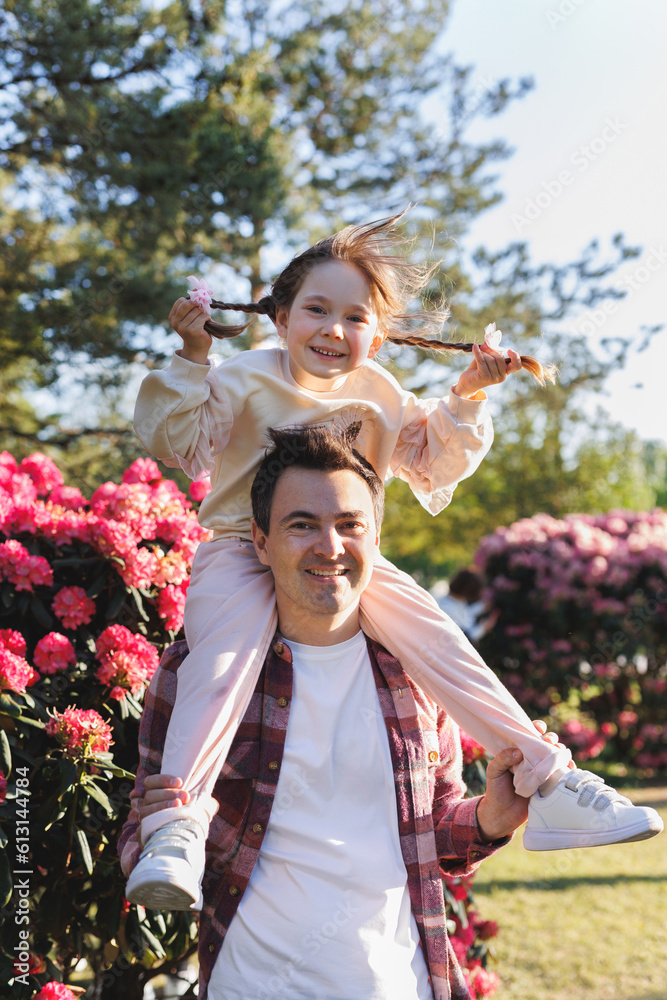 Fototapeta premium Young father and his little girl riding on his shoulders in a blooming park.