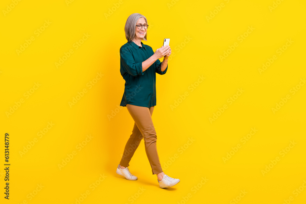 Full size profile portrait of positive lady use smart phone walking isolated on yellow color background Full size profile portrait of positive lady use smart phone walking isolated on yellow color background