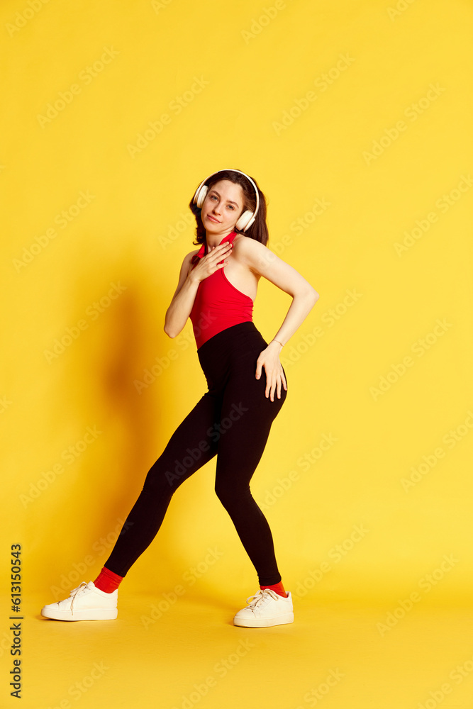 Full-length portrait of young beautiful woman in casual clothes listening to music in headphones and dancing against yellow studio background. Concept of youth, human emotions, lifestyle, ad