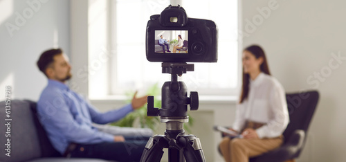 Closeup shot of modern digital video camera in TV studio recording interview with man guest and woman host communicating in blurry background. Banner. Television, mass media, TV show concept