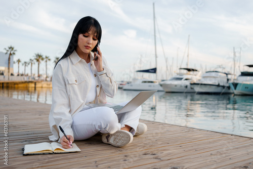 Woman works with a laptop, notepad and talks on the phone sitting in front of the sea (ID: 613163191)