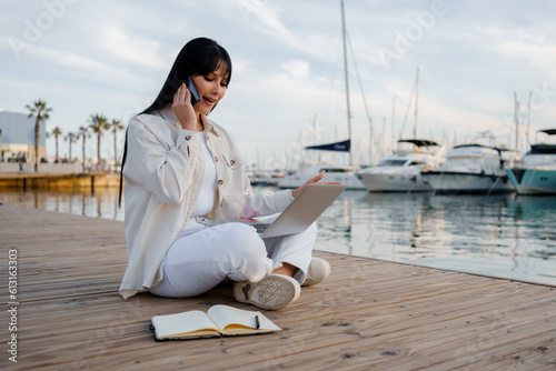 Woman works with a laptop, notepad and talks on the phone sitting in front of the sea (ID: 613163303)