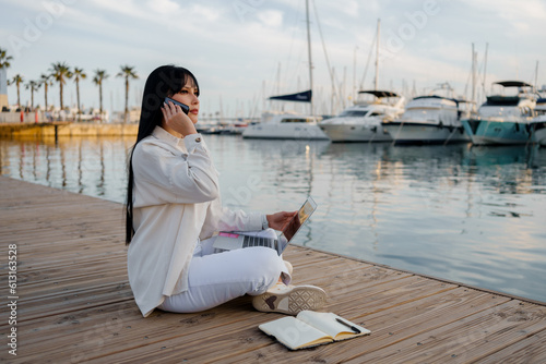 Woman works with a laptop, notepad and talks on the phone sitting in front of the sea (ID: 613163528)