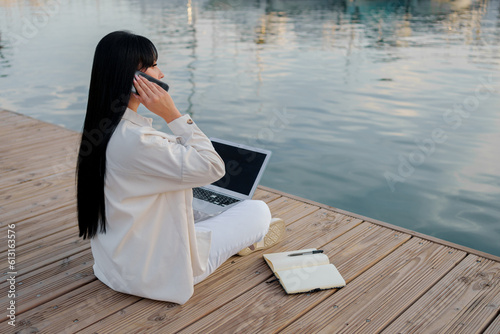 Woman works with a laptop, notepad and talks on the phone sitting in front of the sea (ID: 613163576)