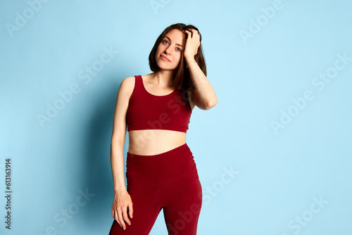 Fototapeta Naklejka Na Ścianę i Meble -  Young brunette woman with slim, fit body posing in red top and leggings, showing expression of disagreement against blue studio background. Concept of youth, human emotions, lifestyle, ad