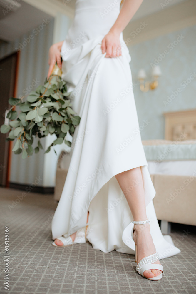 Bride standing indoor in long wedding dress with flowers in hand. Female lift fabric and show shoe adorned with pearls.