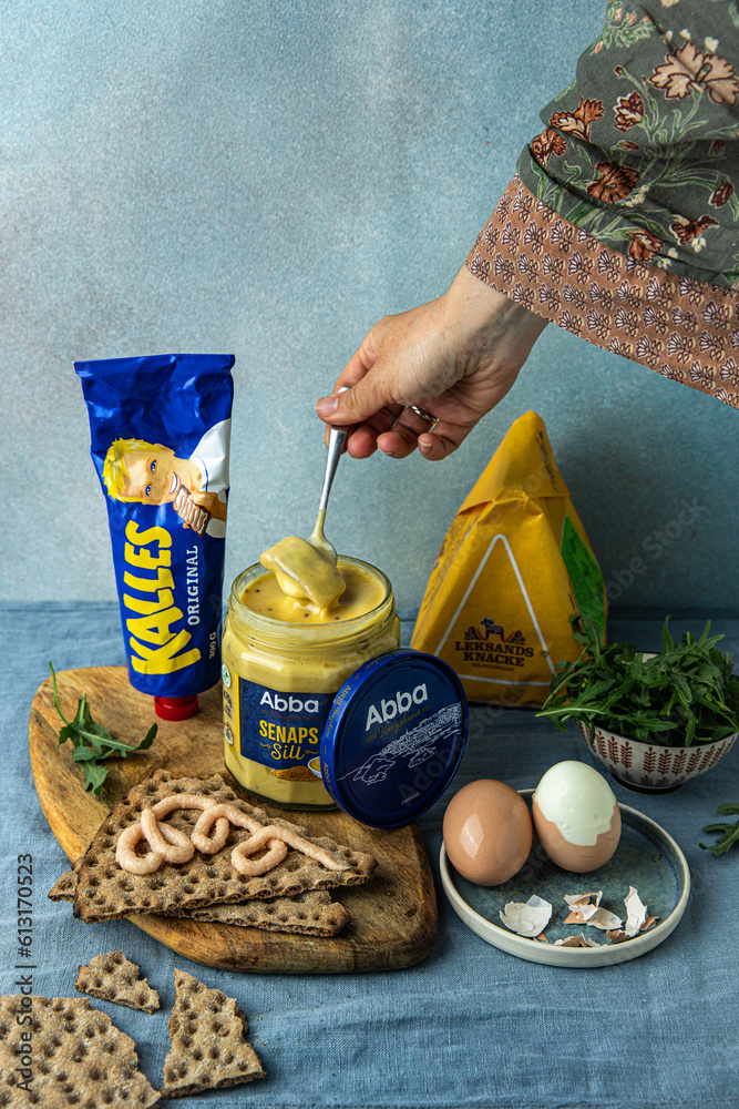 Swedish food. Woman taking out Abba Seafood pickled herring in mustard,  Kalles kaviar in tube and over traditional crisp bread Leksands Knackebrod,  boiled eggs, fresh arugula on light blue background. Stock-Foto