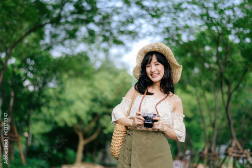 Portrait of asian young woman traveler with weaving hat and basket and a camera on green public park nature background. Journey trip lifestyle, world travel explorer or Asia summer tourism concept. © Jirawatfoto