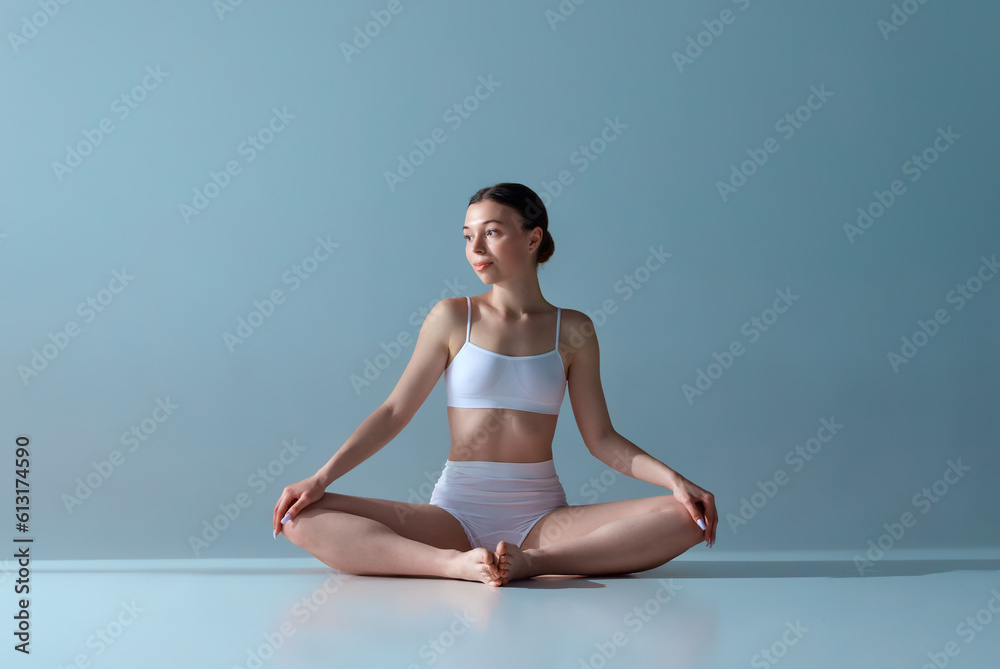 Portrait of beautiful, relaxed, young girl with slim body sitting in lotus pose in underwear against blue studio background Portrait of beautiful, relaxed, young girl with slim body sitting in lotus pose in underwear against blue studio background