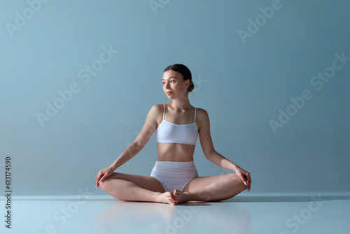 Portrait of beautiful, relaxed, young girl with slim body sitting in lotus pose in underwear against blue studio background