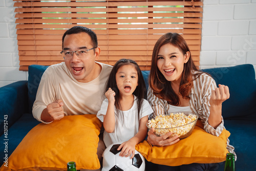 Asian Family with daughter watching sport football cheering excited eating popcorn on sofa in living room at home. enjoy relaxing happiness.