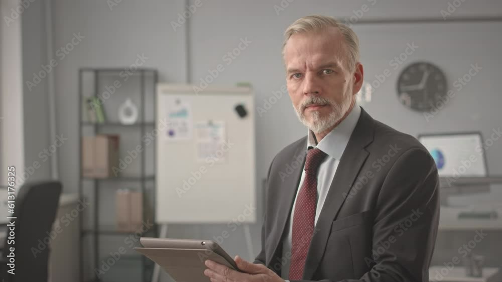 Tilt up portrait of successful middle aged Caucasian businessman in elegant formal suit posing for camera with digital table in hands sitting on work desk in modern office Tilt up portrait of successful middle aged Caucasian businessman in elegant formal suit posing for camera with digital table in hands sitting on work desk in modern office