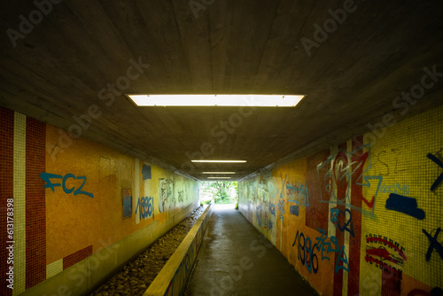 Neon lit urban pedestrian tunnel with graffiti tile side walls and small running water stream, no people