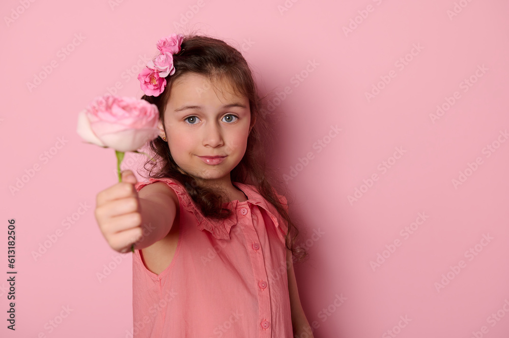 Beautiful little child girl in stylish dress and flowers in hair, showing at camera a beautiful rose flower, smiling over isolated pink background. Happy Mother's Day. International Women's Day Beautiful little child girl in stylish dress and flowers in hair, showing at camera a beautiful rose flower, smiling over isolated pink background. Happy Mother's Day. International Women's Day