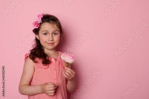 Caucasian lovely child girl, adorable daughter in stylish pink dress, holding a beautiful rose flower, smiling on isolated pink background with copy space. Children. Childhood. Fashionable kids.
