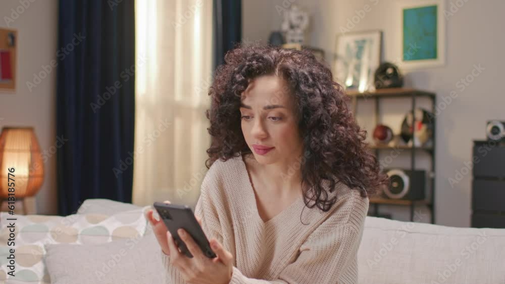 Portrait of Caucasian woman with curly hair holding phone while sitting on soft couch. Beautiful female scrolling news of day or messaging with someone. Having good time at home. Concept technology. Portrait of Caucasian woman with curly hair holding phone while sitting on soft couch. Beautiful female scrolling news of day or messaging with someone. Having good time at home. Concept technology.