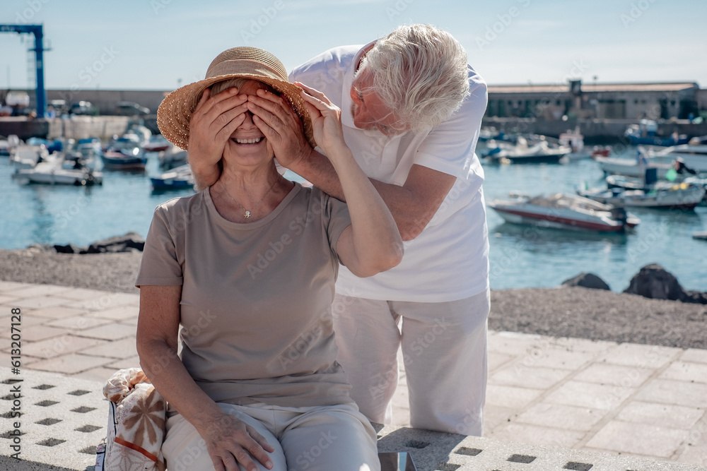 Cheerful smiling elderly family couple sitting on the bench near the sea. Elderly man closes his wife's eyes with his hands. Harbor and boats in the background