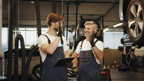 Car service station. Mechanic man shows report on a tablet to the caucasian coworker at garage, A man mechanic and his son discussing repairs done vehicle. Car service, repair, maintenance.