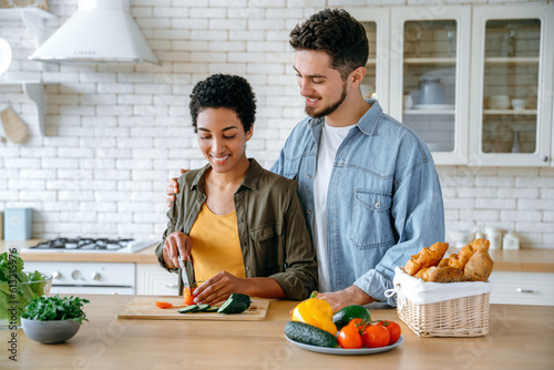 Happy couple in love, dressed in casual wear, young spouses, stand at home in the kitchen, prepare salad together, woman cuts vegetables, man hugs her, they glad to spend time together, smile