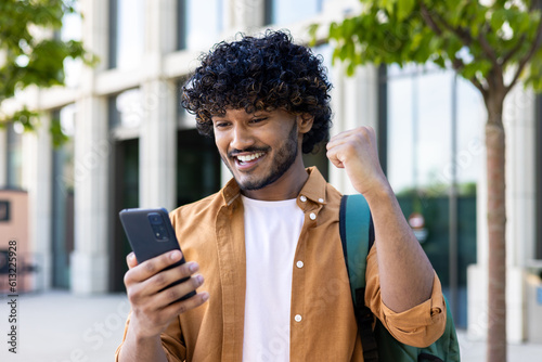 Young hispanic student celebrating victory and happy good news online, man reading page interent received notification and message, hold phone, use app win and good achievement
