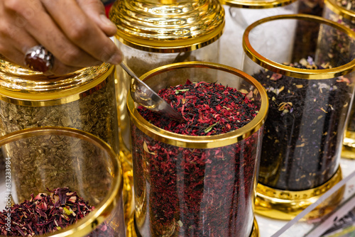 United Arab Emirates, Dubai, April 2023: Spice market, black tea with fruit aroma. Man with spoon takes some tea from the jar. Black fruit tea, spices and herbs stall at the old souk. photo