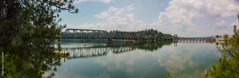 Panorama of metal train bridge over river with a dam and reflections on the water