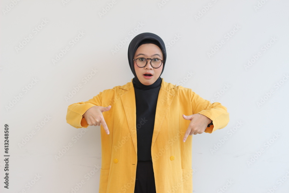 Beautiful young Asian Muslim woman in glasses, hijab and wearing yellow blazer shocked expression with both hands pointing down Beautiful young Asian Muslim woman in glasses, hijab and wearing yellow blazer shocked expression with both hands pointing down