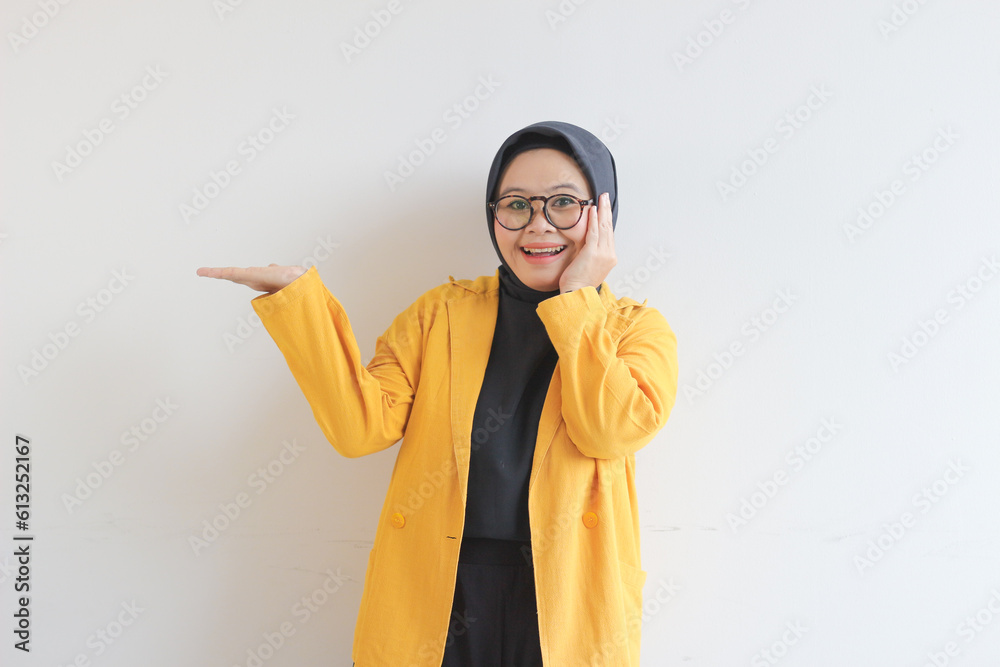 Beautiful young Asian Muslim woman in glasses, hijab and wearing yellow blazer smiling confident and happy with both hands presenting empty space Beautiful young Asian Muslim woman in glasses, hijab and wearing yellow blazer smiling confident and happy with both hands presenting empty space