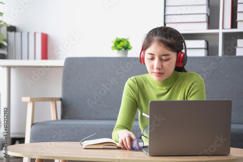 Asian woman with a beautiful face in a green long-sleeve shirt sitting and relaxing watching movies Listening to music on a laptop, mobile phone, and headphones happily inside living room at home.