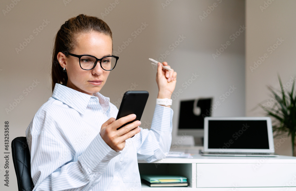 Serious young businesswoman wearing glasses sitting at the desk in her own office space and using her smart phone.
