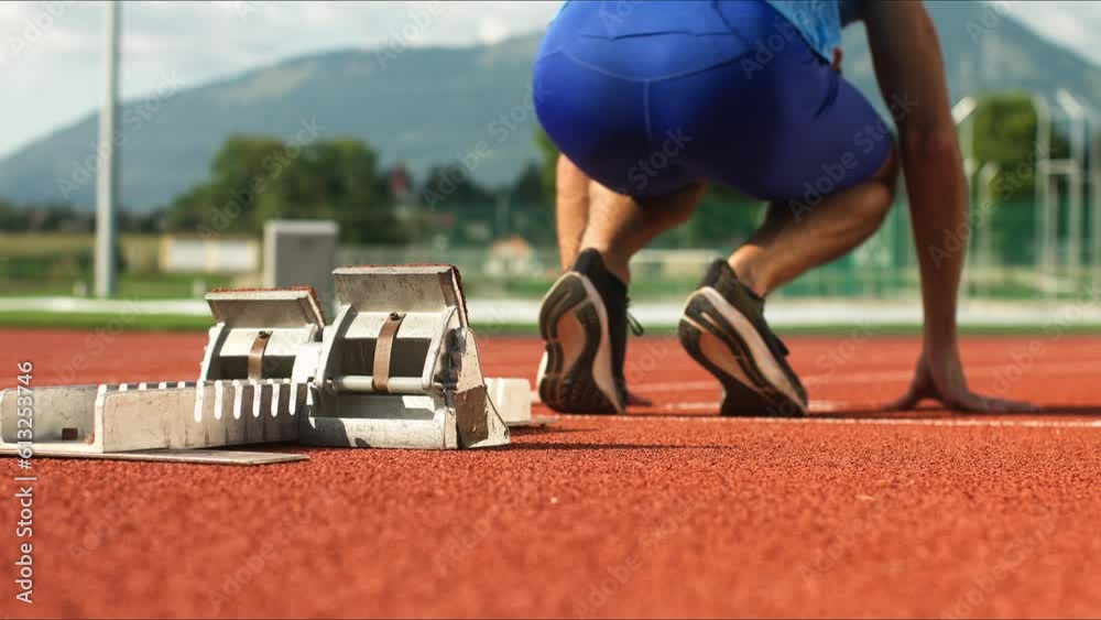 Rear view of a male Caucasian sprinter assuming a starting position in the starting block during running training at an athletic track, ground level close up shot.