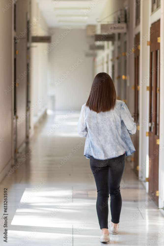female international school student dressed in jeans walked inside school building on corridor in front of classroom alone. back of female student who was walking alone in corridor of school building.