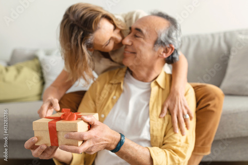 Loving senior wife giving birthday gift box to happy husband and kissing man in cheek, selective focus on present