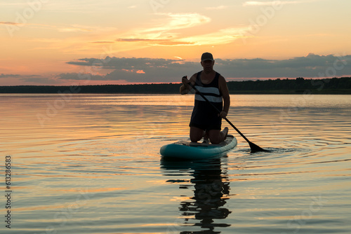 A man on his knees on a SUP board with an oar at sunset swims in the water of the lake against a pink sky reflection in the water. © finist_4