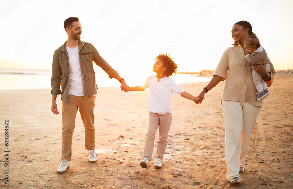 Multiracial parents walking on beach with two sons, holding hands and smiling, enjoying evening by seaside, full length Multiracial parents walking on beach with two sons, holding hands and smiling, enjoying evening by seaside, full length