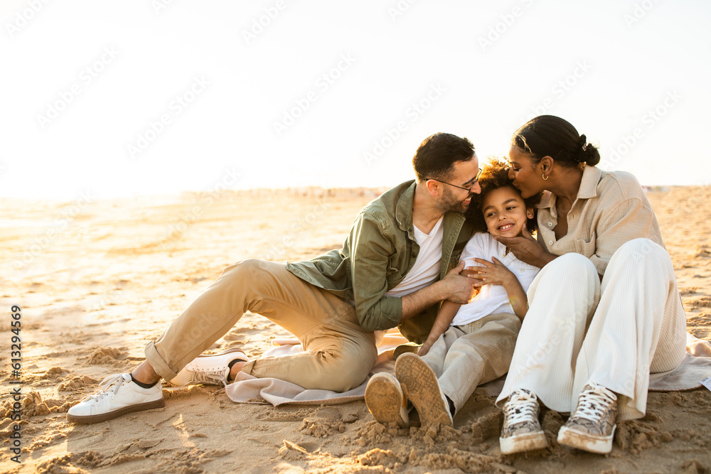 Happy multiracial parents embracing and kissing son while sitting and resting on beach by seaside at sunset