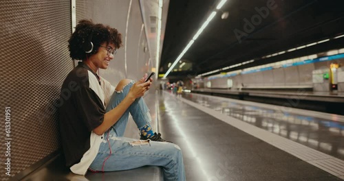Beautiful African-American girl sitting in the subway, listening to music, chatting on her smartphone and waiting for her train. A young black woman is waiting for her train in the subway.