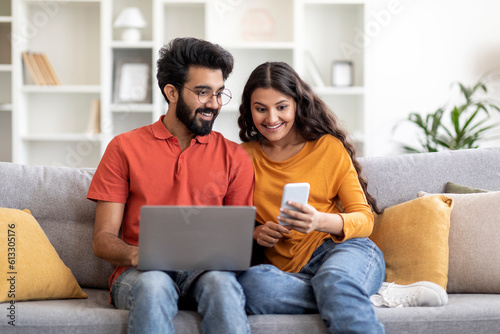 Excited Indian Couple Reading Message On Smartphone And Using Laptop At Home