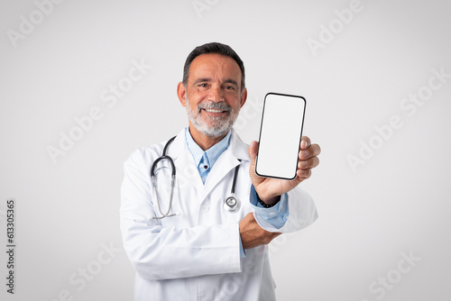 Cheerful caucasian mature man doctor in white coat shows smartphone with blank screen