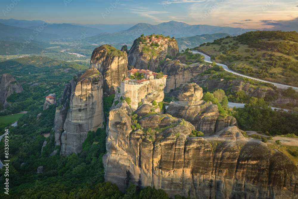 Aerial view of Meteora Monastery complex on rocks, illuminated by rising sun against mountains in background. UNESCO World Heritage Site. Greece.