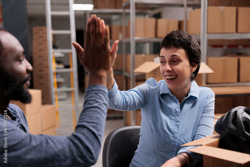 Smiling employees doing high five in storehouse after finishing putting helmet order for client in cardboard boxe, discussing about shipping logistics. Happy team celebrating triumph in warehouse Smiling employees doing high five in storehouse after finishing putting helmet order for client in cardboard boxe, discussing about shipping logistics. Happy team celebrating triumph in warehouse