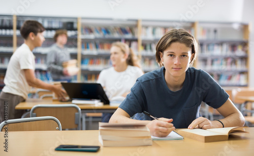 Focused fifteen-year-old schoolboy sitting at a desk in the library is writing an synopsis from a textbook