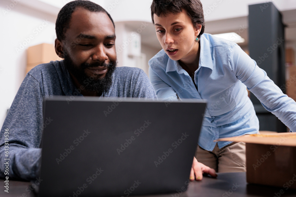 Diverse team sitting at table in storehouse checking shipping logistics on laptop computer before start delivering trendy clothes for clients. Employees working with carton boxes in fashion warehouse Diverse team sitting at table in storehouse checking shipping logistics on laptop computer before start delivering trendy clothes for clients. Employees working with carton boxes in fashion warehouse
