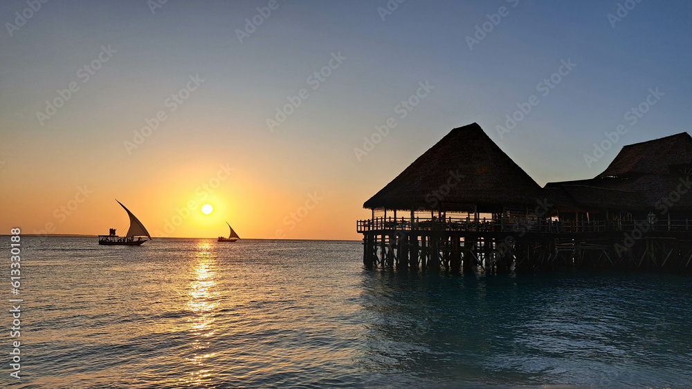 Kendwa, Zanzibar Island, Tanzania dhow boats sailing and a wooden thatched bungalow pier against the setting sun and cloudy sky. - obrazy, fototapety, plakaty 