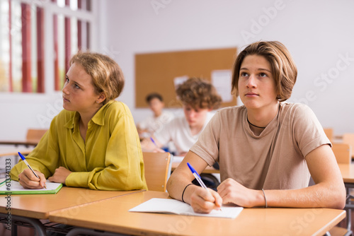 Teenage boy and girl listening to teacher and writing exercises in notebook at lesson in secondary school