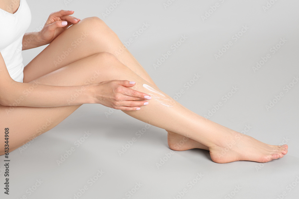 Woman applying body cream onto her smooth legs on light grey background, closeup