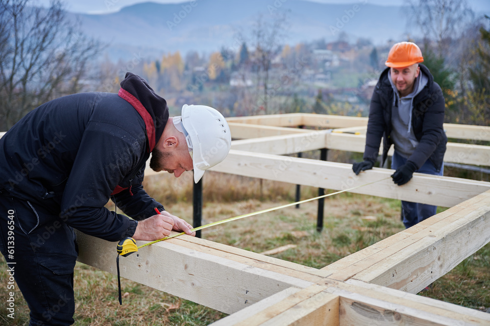 Man worker building wooden frame house on pile foundation. Carpenter using tape measure for measuring wooden planks and making marks with pencil. Carpentry concept. Man worker building wooden frame house on pile foundation. Carpenter using tape measure for measuring wooden planks and making marks with pencil. Carpentry concept.